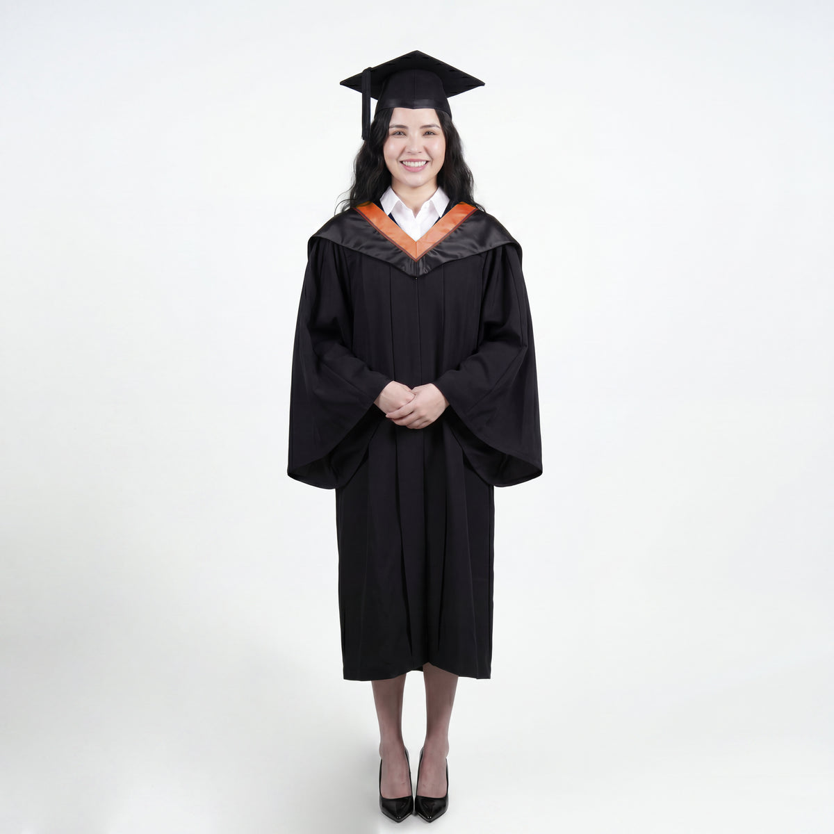 A woman wearing a black graduation gown and matching mortarboard cap stands against a light gray studio background.
The gown has a wide collar with copper and brown trim accent hood, and the model is posed with hands clasped in front.