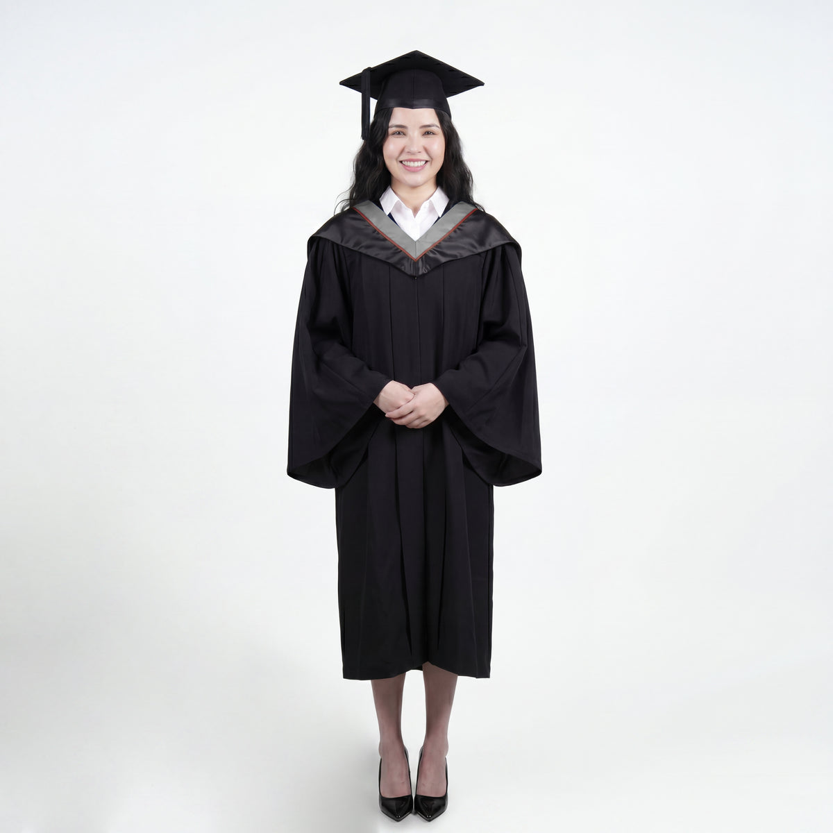 A woman wearing a black graduation gown and matching mortarboard cap stands against a light gray studio background.
The gown has a wide collar with grey and brown trim accent hood, and the model is posed with hands clasped in front.