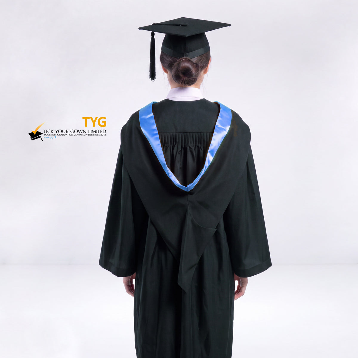 A back view of a woman wearing a black graduation gown and a black mortarboard cap stands against a light gray studio background.
The gown has a light blue bachelor hood, and a white collared shirt is visible underneath.