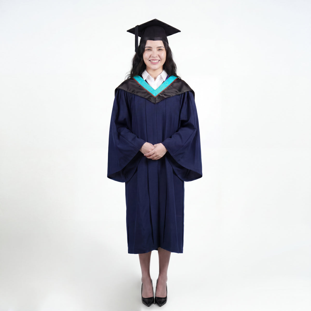 A woman wearing a black graduation gown and matching mortarboard cap stands against a light gray studio background.
The gown has a wide collar with light blue and brown trim accent hood, and the model is posed with hands clasped in front.