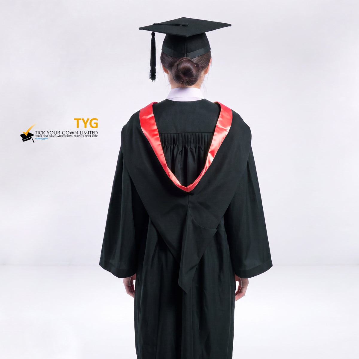 A back view of a woman wearing a black graduation gown and a black mortarboard cap stands against a light gray studio background.
The gown has a red bachelor hood, and a white collared shirt is visible underneath.