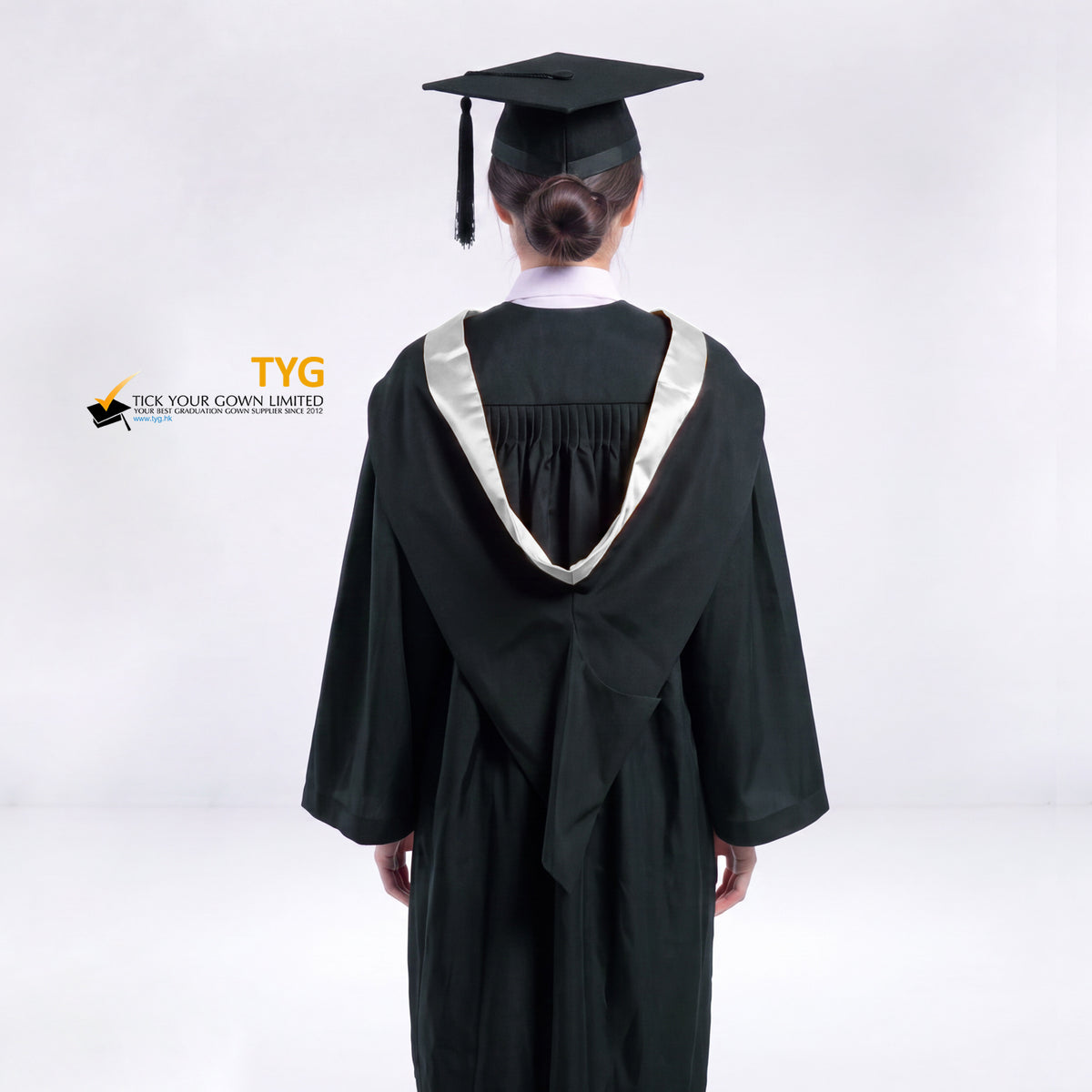 A back view of a woman wearing a black graduation gown and a black mortarboard cap stands against a light gray studio background.
The gown has a silver bachelor hood, and a white collared shirt is visible underneath.