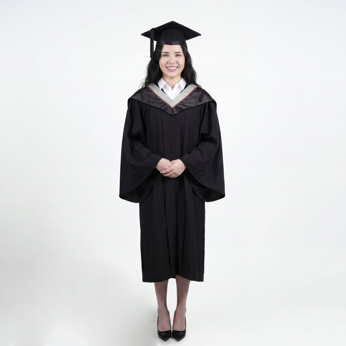 A woman wearing a black graduation gown and matching mortarboard cap stands against a light gray studio background.
The gown has a wide collar with silver and brown trim accent hood, and the model is posed with hands clasped in front.