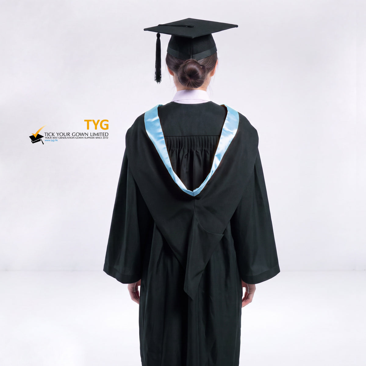 A back view of a woman wearing a black graduation gown and a black mortarboard cap stands against a light gray studio background.
The gown has a sky blue bachelor hood, and a white collared shirt is visible underneath.