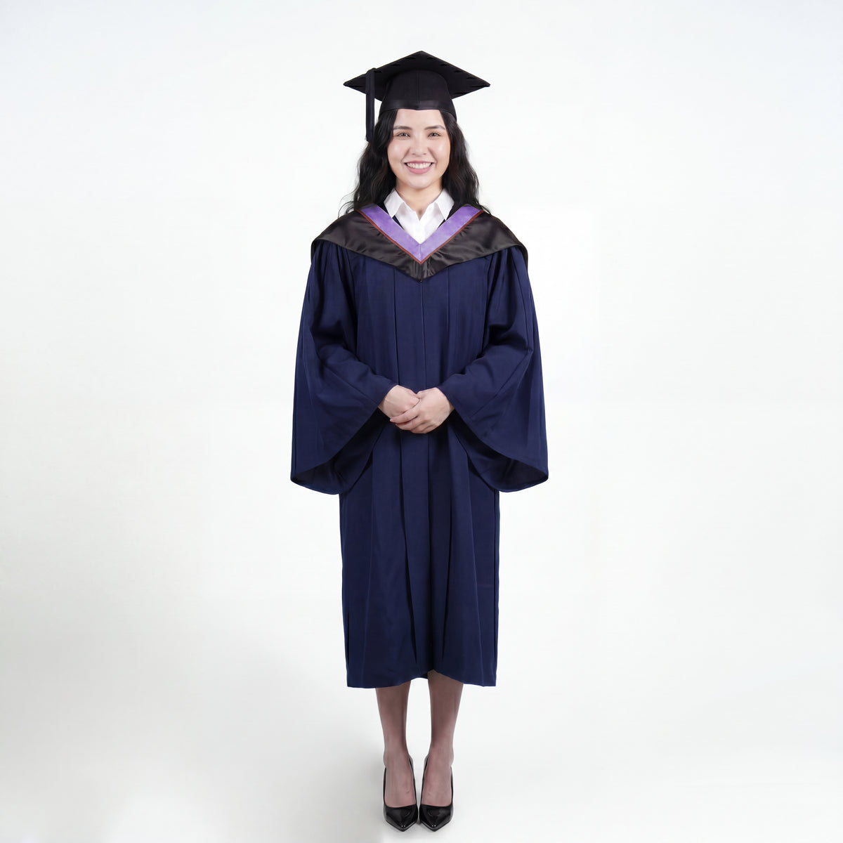 A woman wearing a navy blue graduation gown and matching mortarboard cap stands against a light gray studio background.
The gown has a wide collar with light purple and brown trim accent hood, and the model is posed with hands clasped in front.