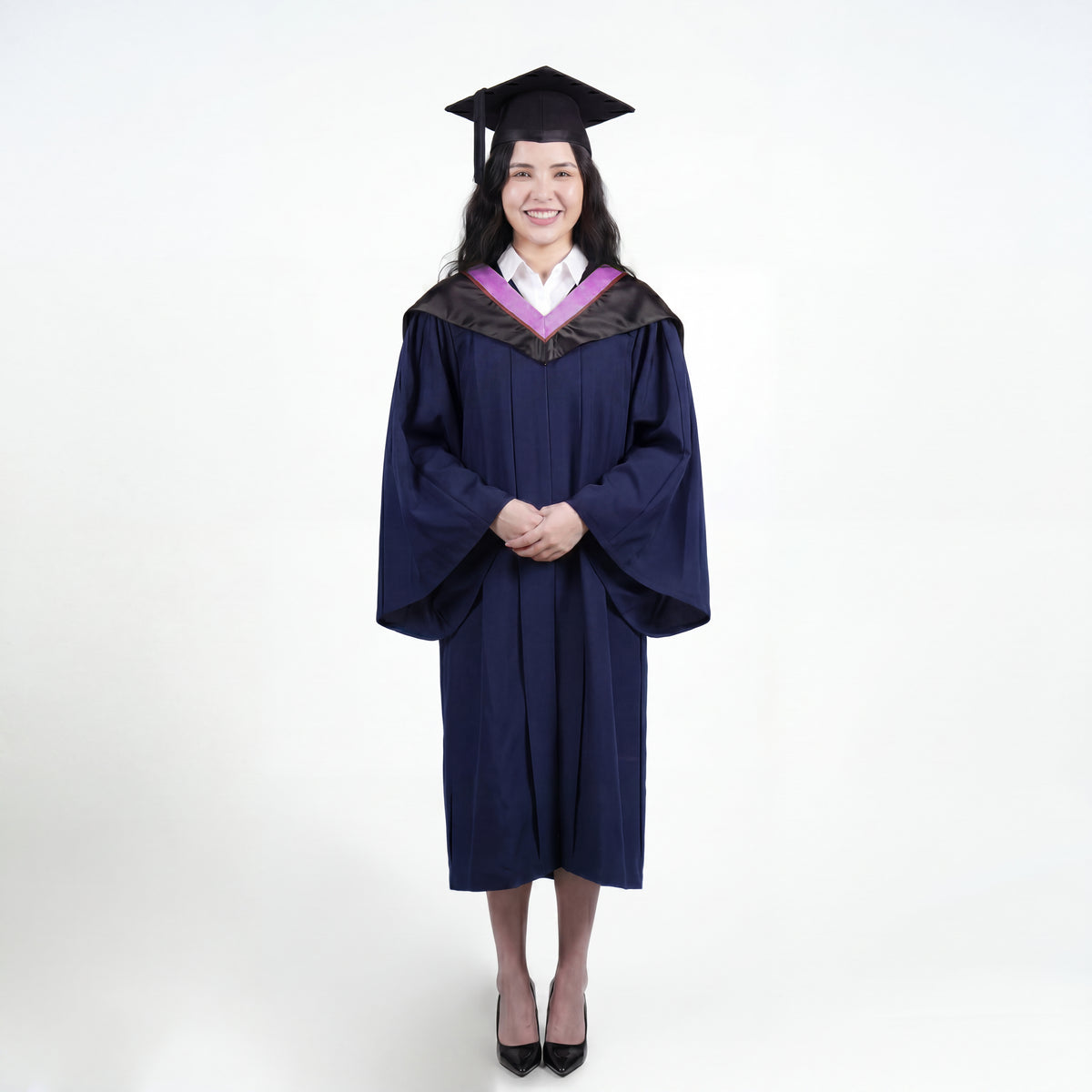 A woman wearing a navy blue graduation gown and matching mortarboard cap stands against a light gray studio background.
The gown has a wide collar with pink and brown trim accent hood, and the model is posed with hands clasped in front.
