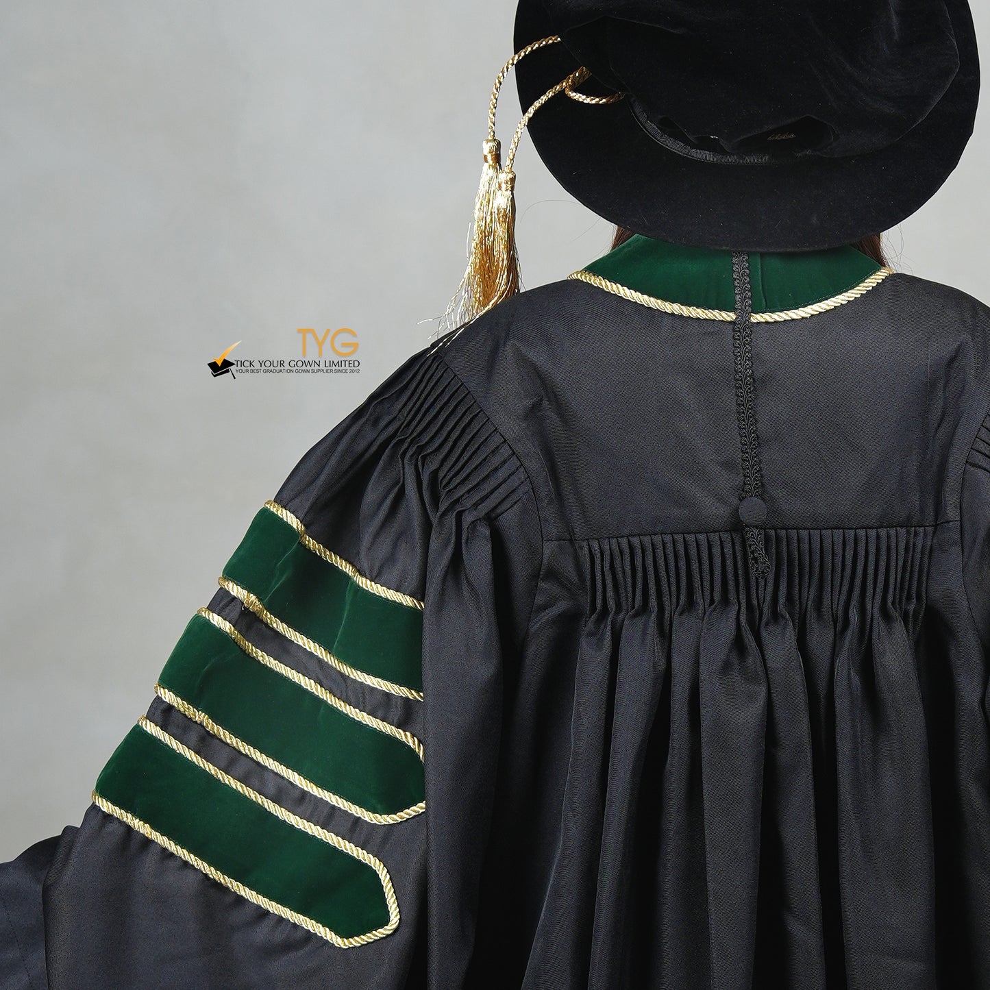 A detailed back view of a person wearing a PhD graduation gown in black with forest green velvet panels, matching sleeve bars, gold trim, and a matching doctoral bonnet with gold tassel.