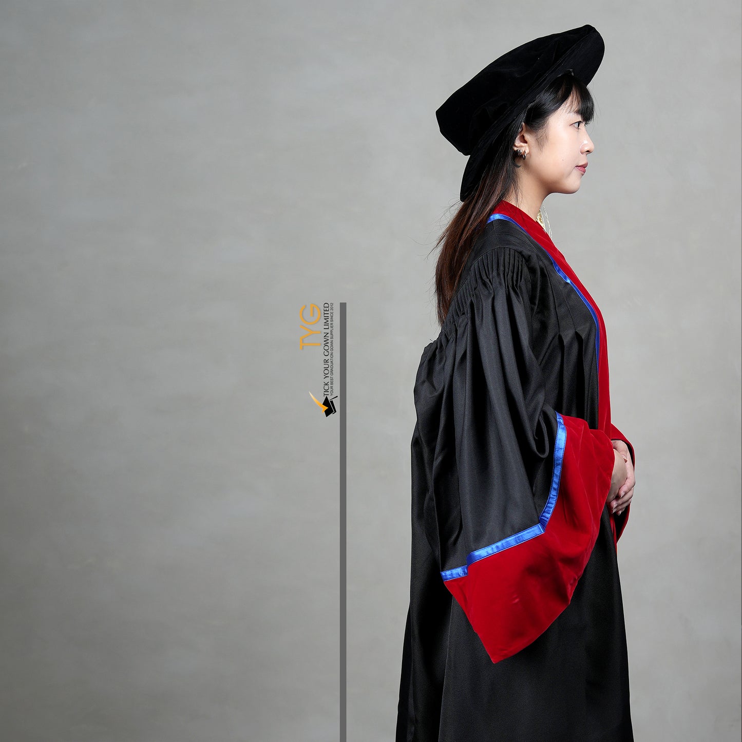 A detailed side view of a person wearing a premium honorary graduation gown in black with red velvet panels, royal blue trim, red velvet sleeve cuffs, and a matching doctoral bonnet with gold tassel.