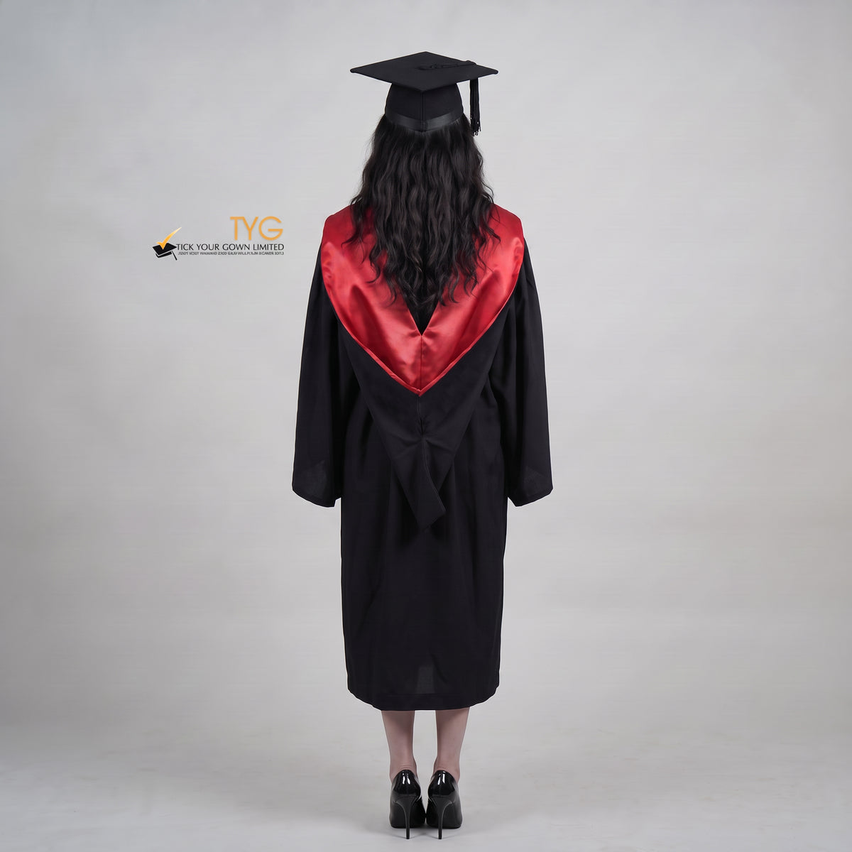A back view woman wearing a University of Edinburgh bachelor graduation gown and a black mortarboard cap stands against a light gray studio background. 

The gown has a red and black draped hood, and she is wearing a white collared shirt underneath.
