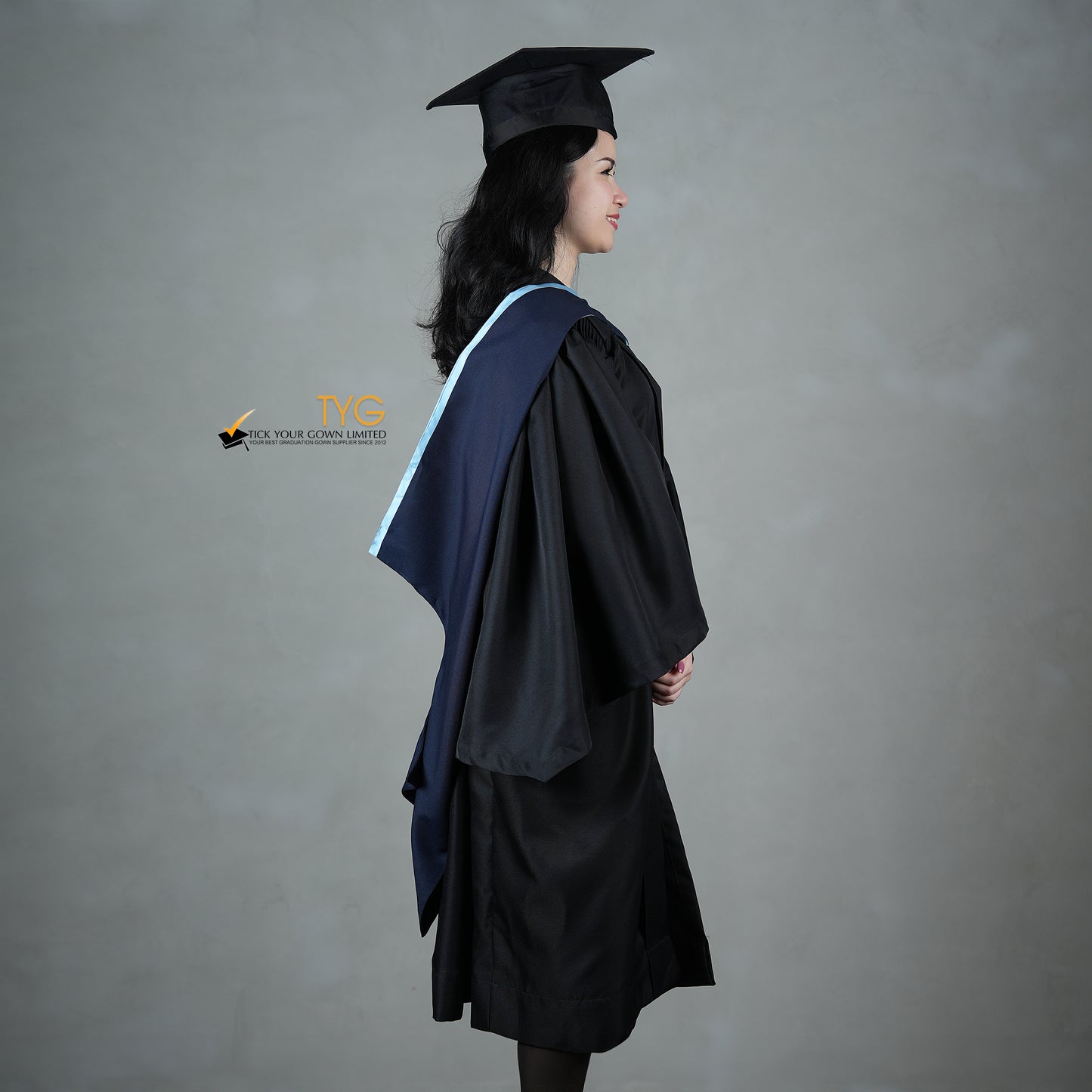 A side view of a graduate in a black cap and gown with a navy blue hood with light blue lining against a gray background