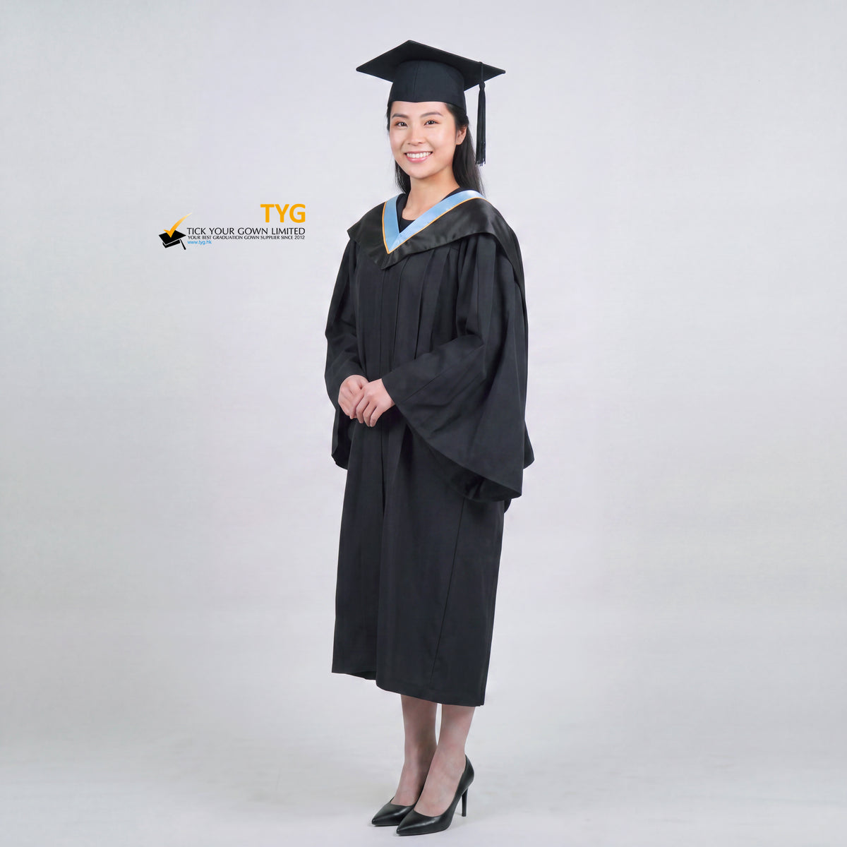 A woman wearing a black graduation gown and matching mortarboard cap stands against a light gray studio background on a side view pose.

The gown has a wide collar with light blue and brown trim accent hood, and the model is posed with hands clasped in front.