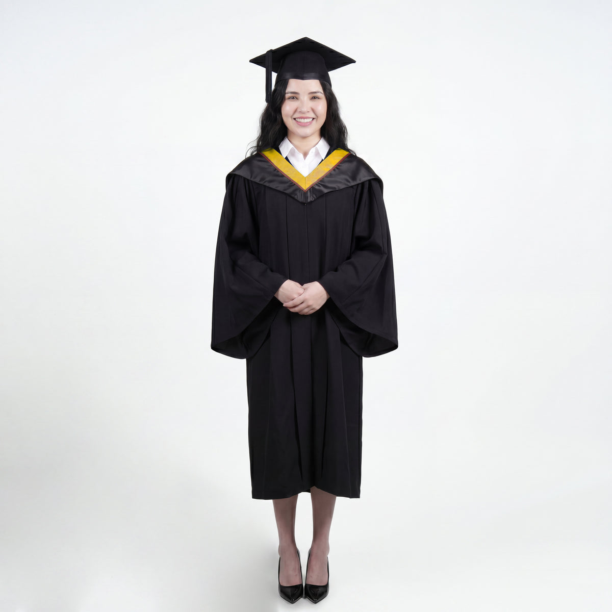 A woman wearing a black graduation gown and matching mortarboard cap stands against a light gray studio background.

The gown has a wide collar with gold and brown trim accent hood, and the model is posed with hands clasped in front.