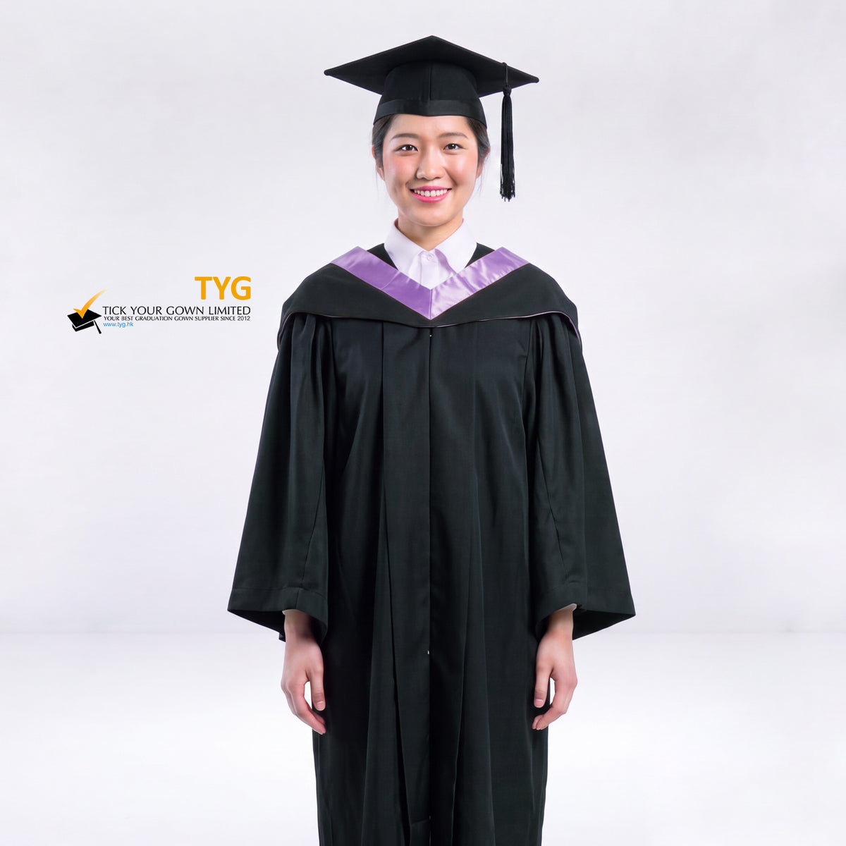 A woman wearing a black graduation gown and a black mortarboard cap stands against a light gray studio background. 

The gown has a lilac bachelor hood, and a white collared shirt is visible underneath.