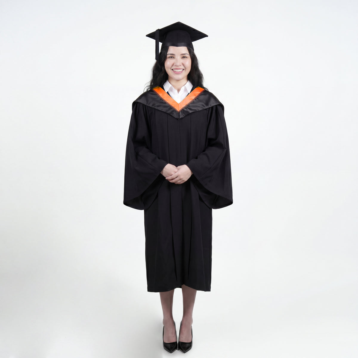 A woman wearing a black graduation gown and matching mortarboard cap stands against a light gray studio background.

The gown has a wide collar with orange and brown trim accent hood, and the model is posed with hands clasped in front.