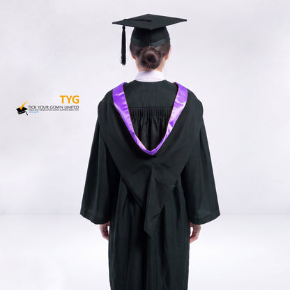 A back view of a woman wearing a black graduation gown and a black mortarboard cap stands against a light gray studio background. 

The gown has a purple bachelor hood, and a white collared shirt is visible underneath.