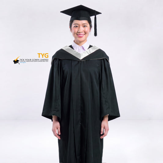 A woman wearing a black graduation gown and a black mortarboard cap stands against a light gray studio background. 

The gown has a silver bachelor hood, and a white collared shirt is visible underneath.