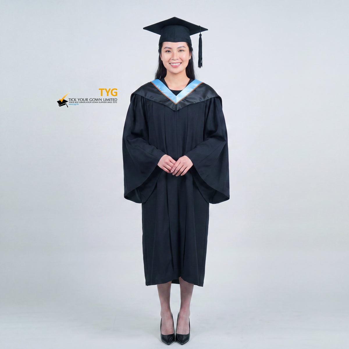 A woman wearing a black graduation gown and matching mortarboard cap stands against a light gray studio background.

The gown has a wide collar with light blue and brown trim accent hood, and the model is posed with hands clasped in front.