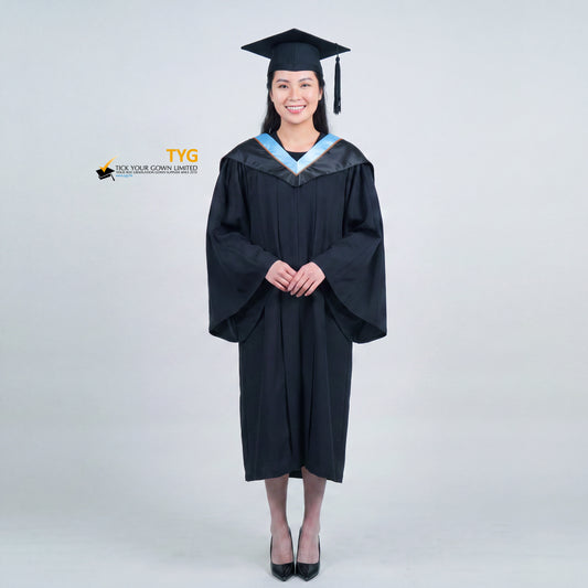 A woman wearing a black graduation gown and matching mortarboard cap stands against a light gray studio background.

The gown has a wide collar with light blue and brown trim accent hood, and the model is posed with hands clasped in front.