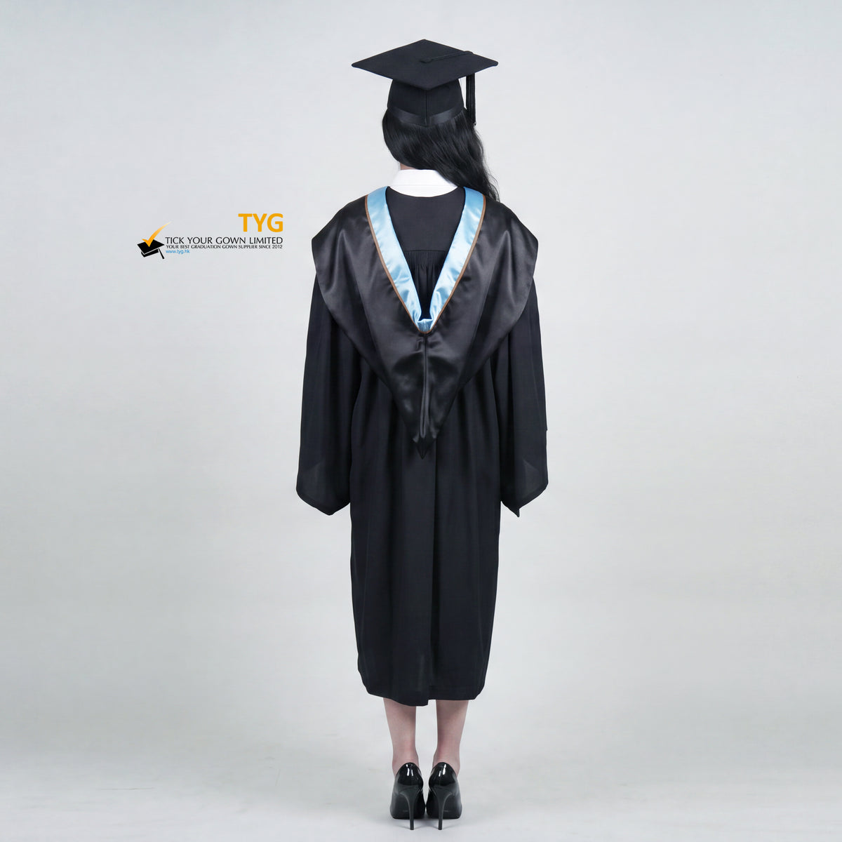 A back view of a woman wearing a black graduation gown and matching mortarboard cap stands against a light gray studio background.

The gown has a wide collar with copper and brown trim accent hood, and the model is posed with hands clasped in front.