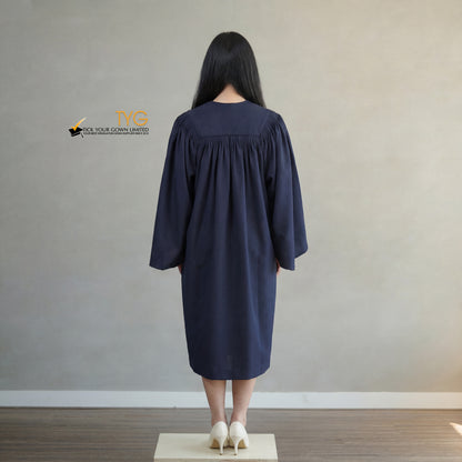 A back view of a person wearing a bachelors graduation gown in navy blue standing on a wooden platform against a plain wall.