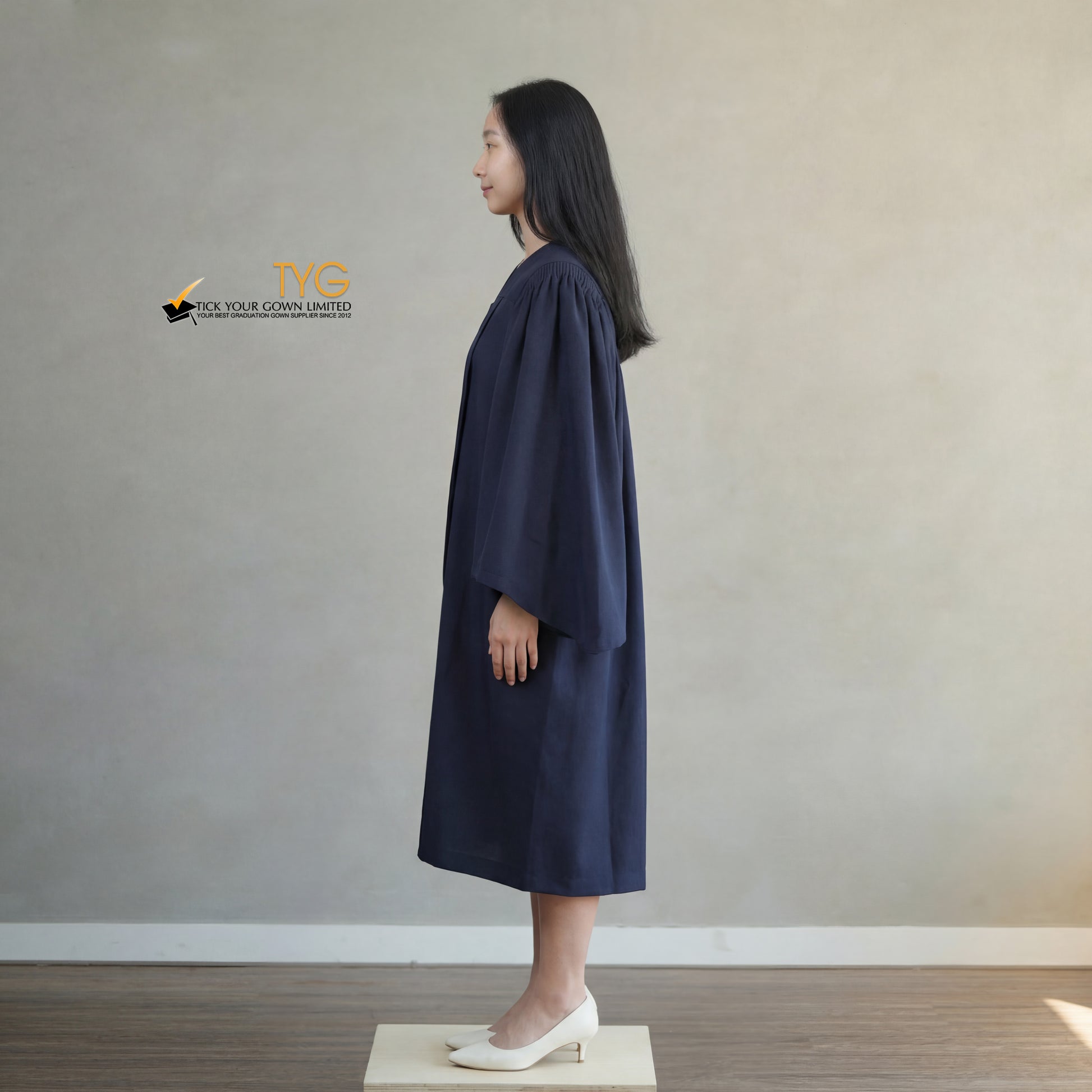 A side view of a person wearing a bachelors graduation gown in navy blue standing on a wooden platform against a plain wall.