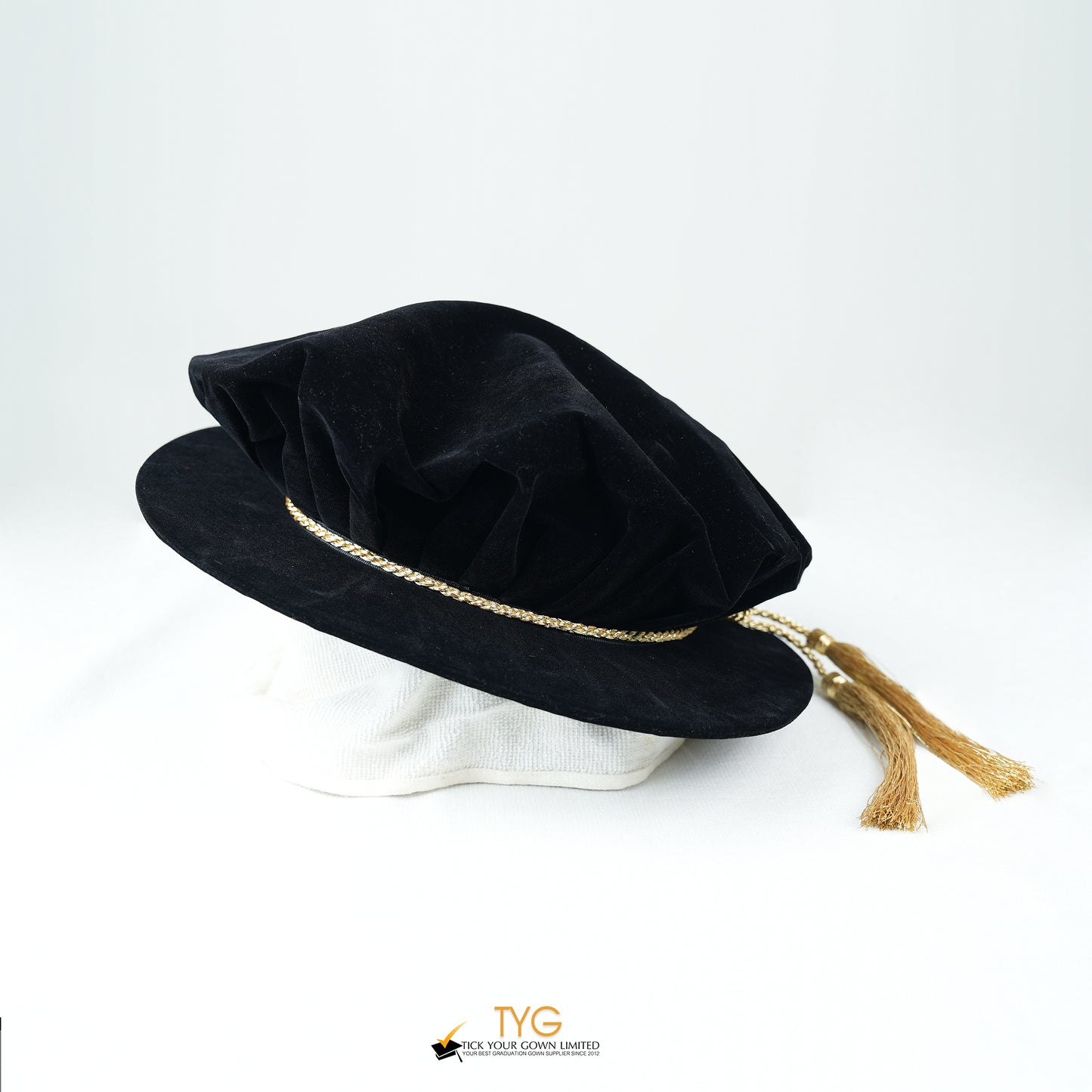 Side view of a black velvet doctoral bonnet featuring a gathered crown, metallic gold braided cord, and gold metallic tassels extending to the right against a plain white background.