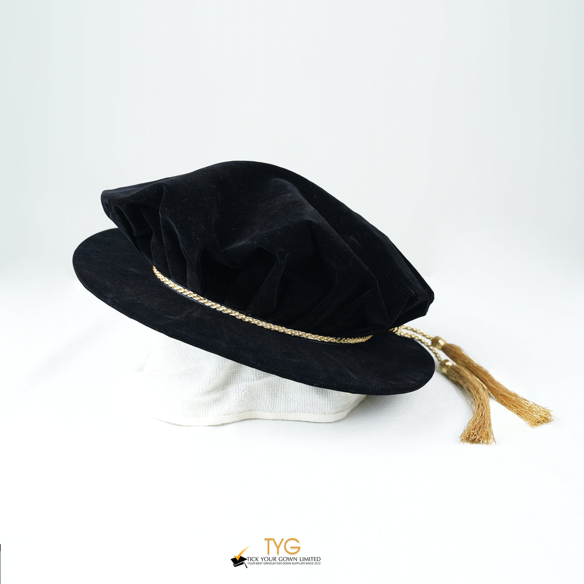 Side view of a black velvet doctoral bonnet featuring a gathered crown, metallic gold braided cord, and gold metallic tassels extending to the right against a plain white background.