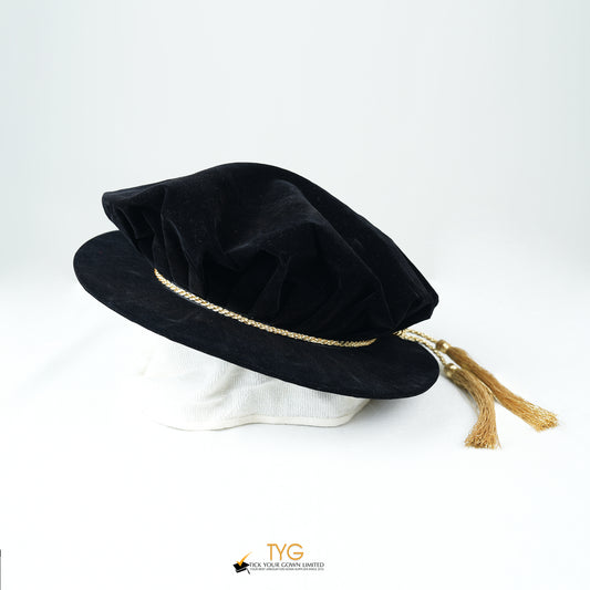 Side view of a black velvet doctoral bonnet featuring a gathered crown, metallic gold braided cord, and gold metallic tassels extending to the right against a plain white background.