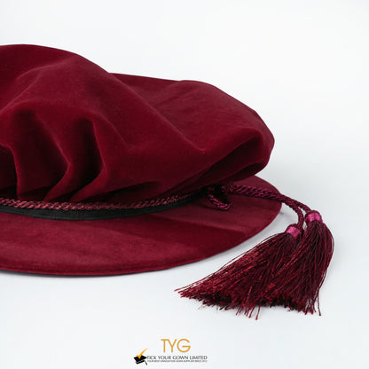 Close-up side view of a maroon velvet doctoral bonnet showing the pleated fabric crown, black band with decorative cord, and draped maroon tassels on a white background.