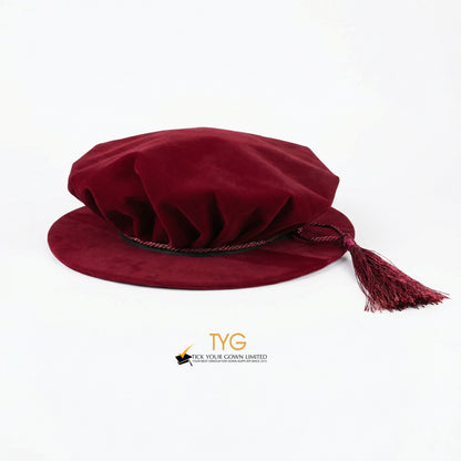 Eye-level front view of a maroon velvet doctoral bonnet showing its soft pleated crown, dark trim, and a draped maroon tassel extending to the right against a plain white background.