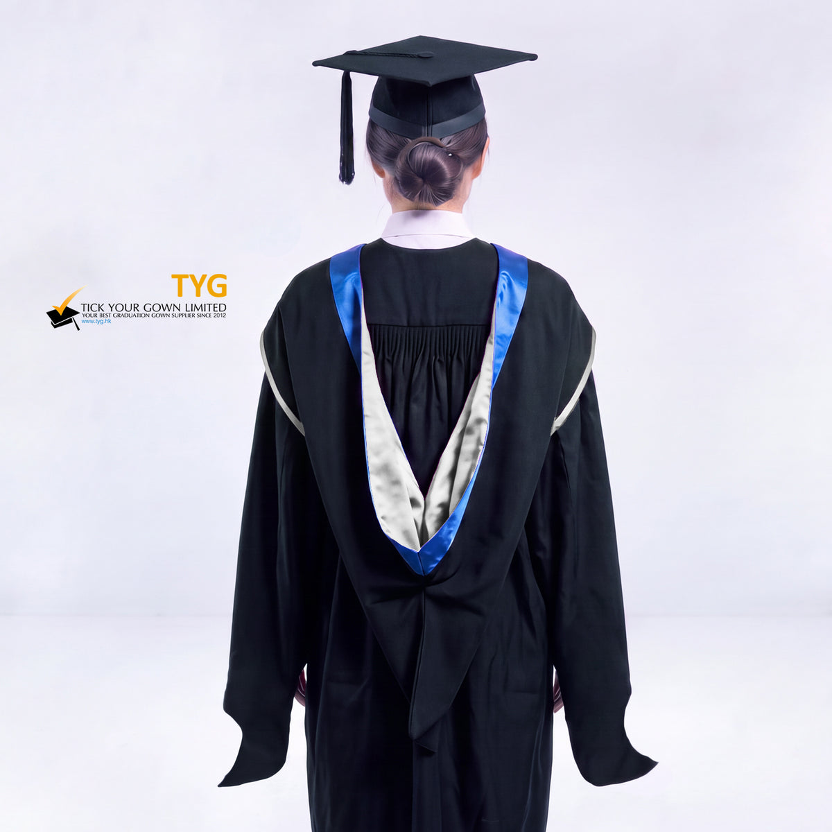 A back view of a woman in a black master's graduation gown classic UK with a blue hood with silver trim accent and with a black mortarboard cap with a black tassel against a gray background.