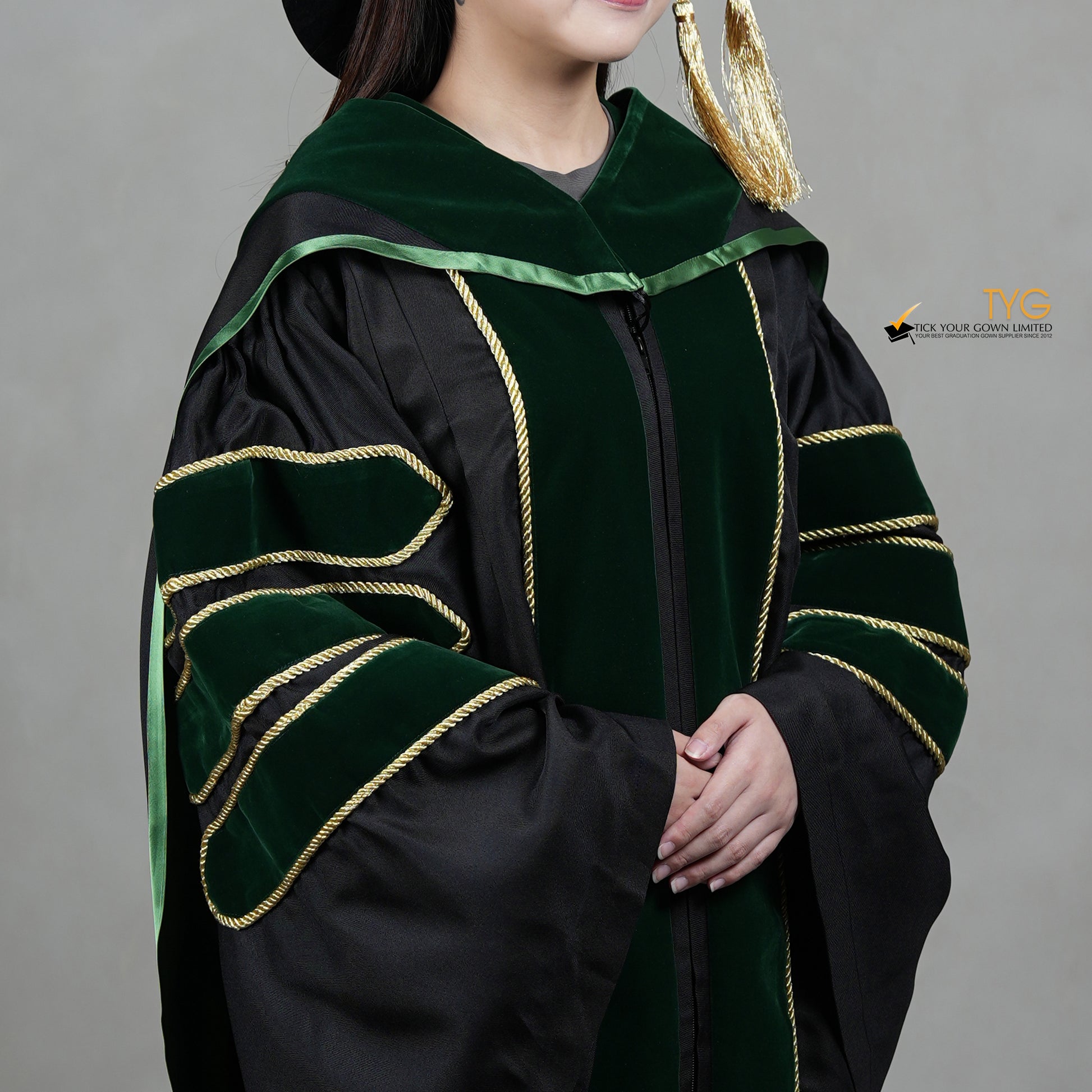 A detailed profile view of a person wearing a PhD graduation gown in black with forest green velvet panels, matching sleeve bars, gold trim, and a matching doctoral bonnet with gold tassel.