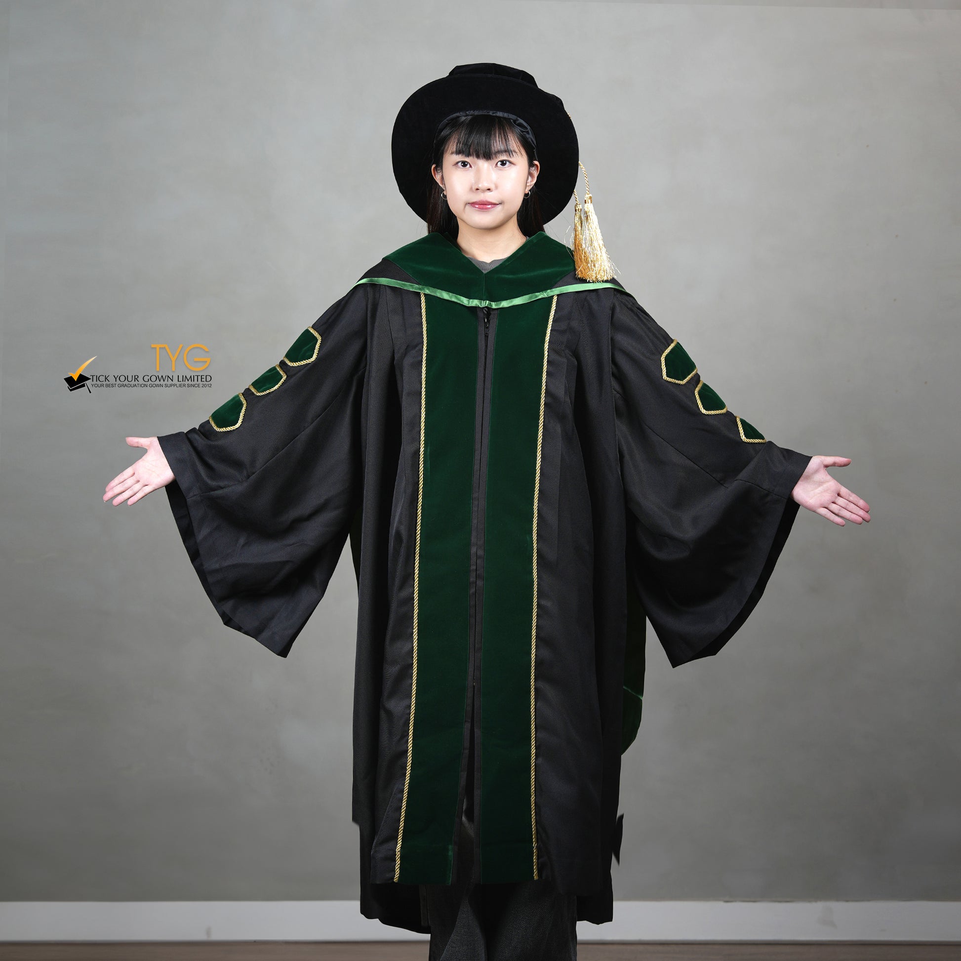 A showcase front view of a person wearing a PhD graduation gown in black with forest green velvet panels, matching sleeve bars, gold trim, and a matching doctoral bonnet with gold tassel.