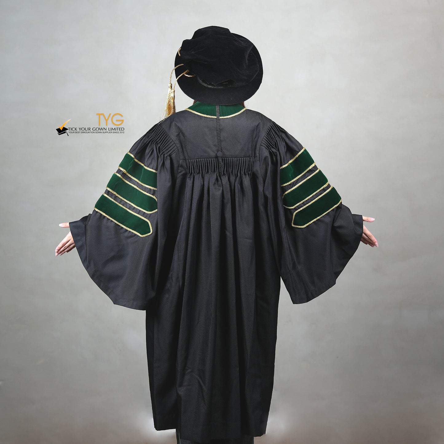 A back view of a person wearing a PhD graduation gown in black with forest green velvet panels, matching sleeve bars, gold trim, and a matching doctoral bonnet with gold tassel.