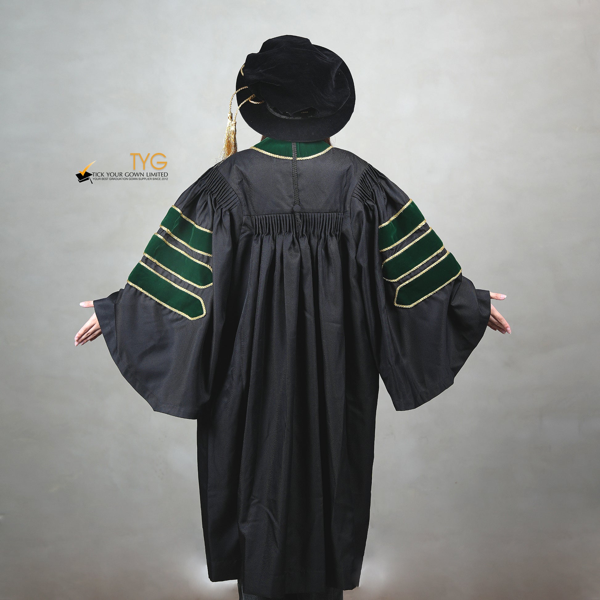 A back view of a person wearing a PhD graduation gown in black with forest green velvet panels, matching sleeve bars, gold trim, and a matching doctoral bonnet with gold tassel.