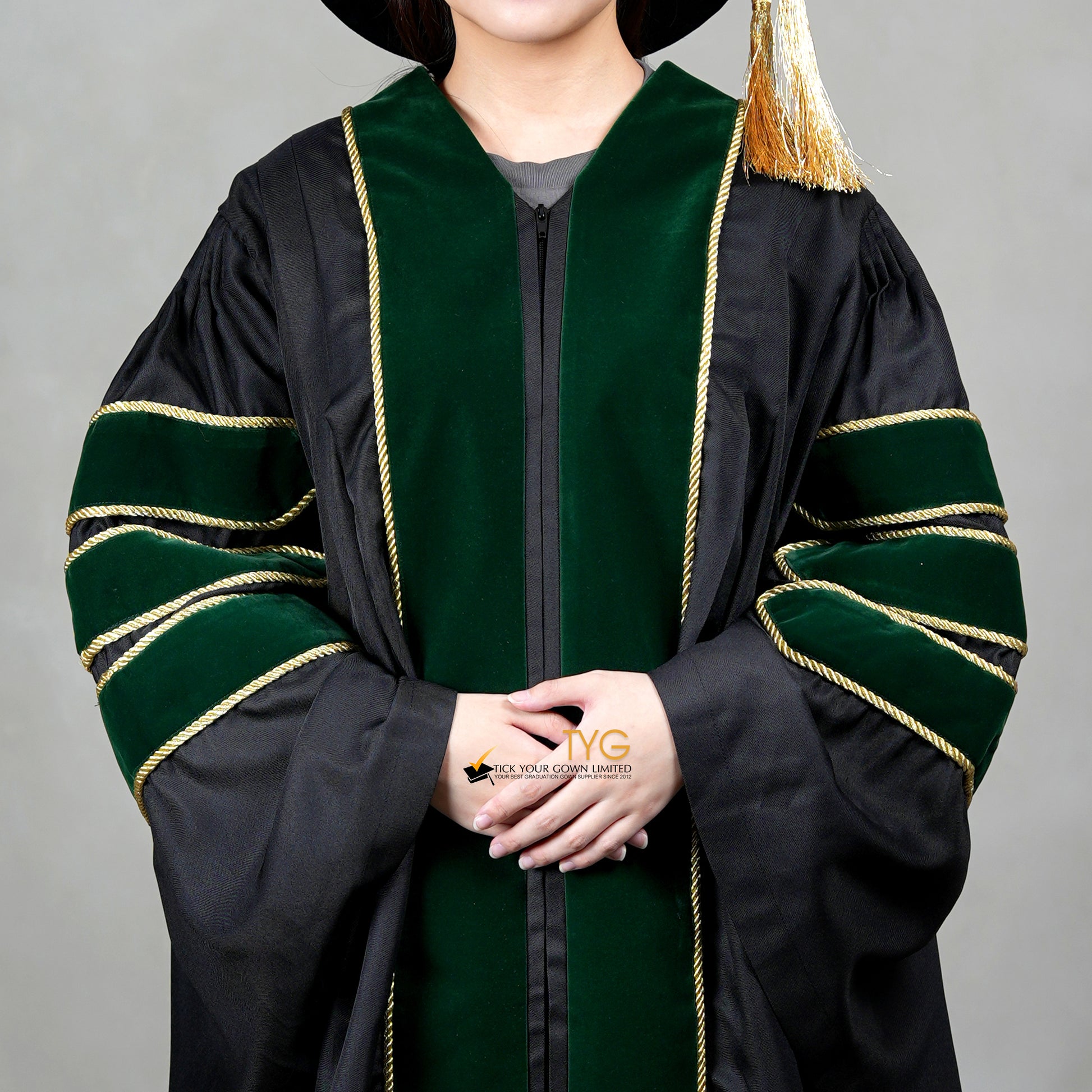 A detailed view of a person wearing a PhD graduation gown in black with forest green velvet panels, matching sleeve bars, gold trim, and a matching doctoral bonnet with gold tassel.