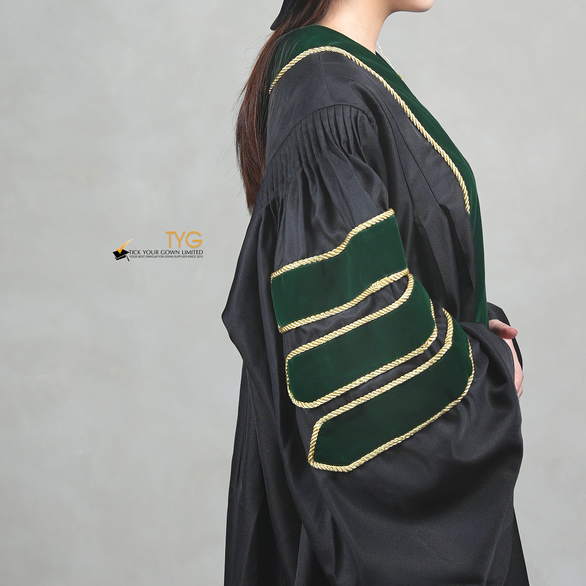 A detailed side view of a person wearing a PhD graduation gown in black with forest green velvet panels, matching sleeve bars, gold trim, and a matching doctoral bonnet with gold tassel.