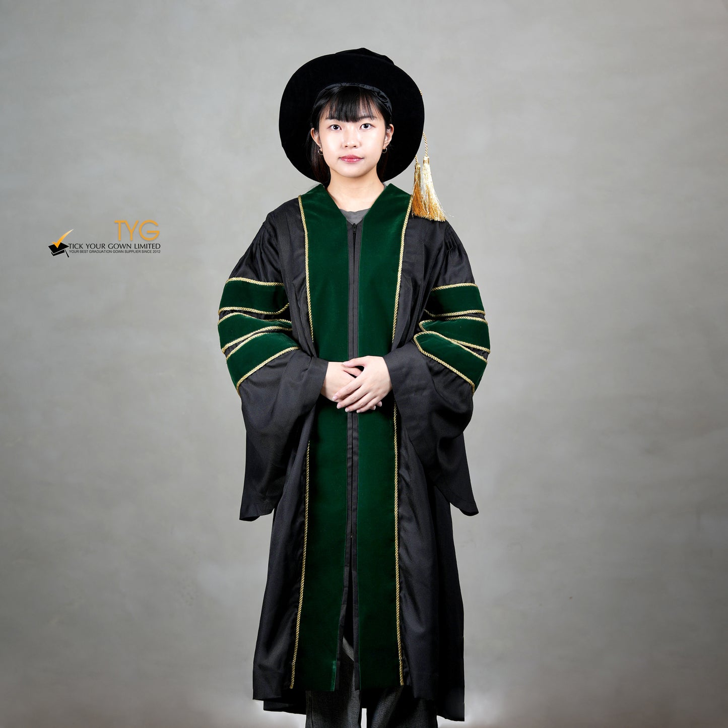 Front view of a person wearing a PhD graduation gown in black with forest green velvet panels, matching sleeve bars, gold trim, and a matching doctoral bonnet with gold tassel.