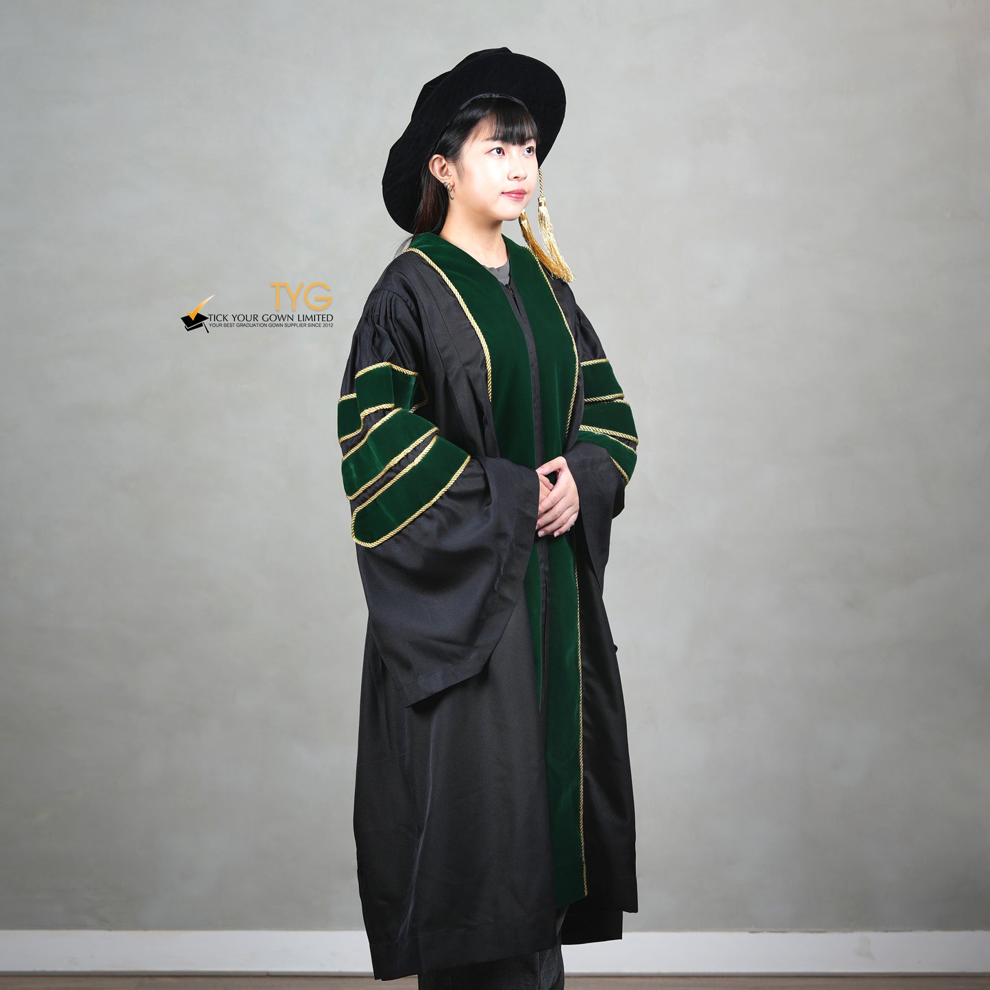 A profile view of a person wearing a PhD graduation gown in black with forest green velvet panels, matching sleeve bars, gold trim, and a matching doctoral bonnet with gold tassel.