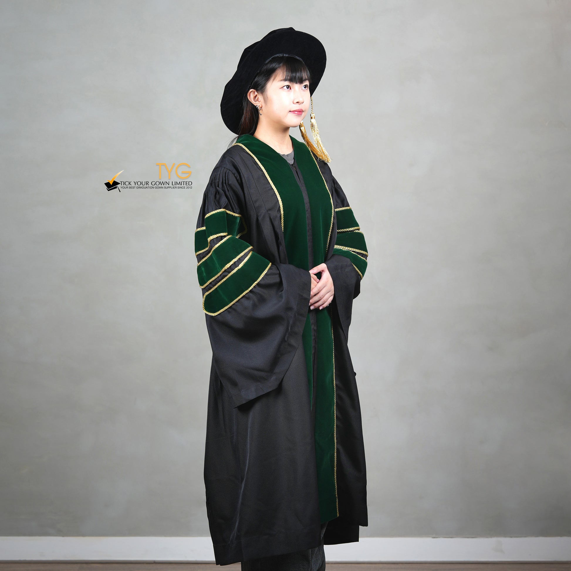 A profile view of a person wearing a PhD graduation gown in black with forest green velvet panels, matching sleeve bars, gold trim, and a matching doctoral bonnet with gold tassel.