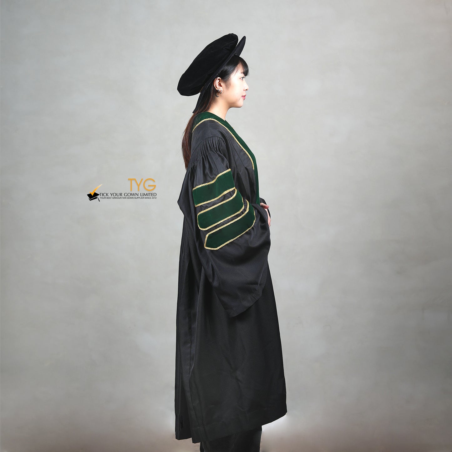 A side view of a person wearing a PhD graduation gown in black with forest green velvet panels, matching sleeve bars, gold trim, and a matching doctoral bonnet with gold tassel.