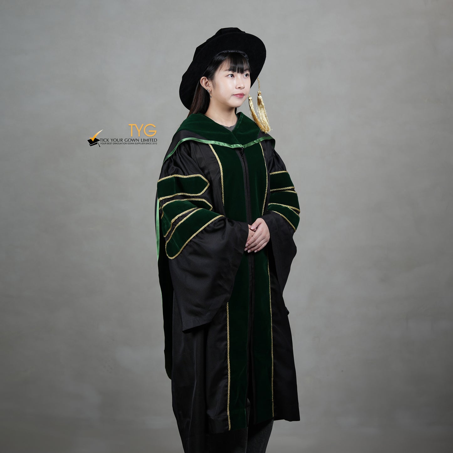 A profile view of a person wearing a PhD graduation gown in black with forest green velvet panels, matching sleeve bars, gold trim, and a matching doctoral bonnet with gold tassel.