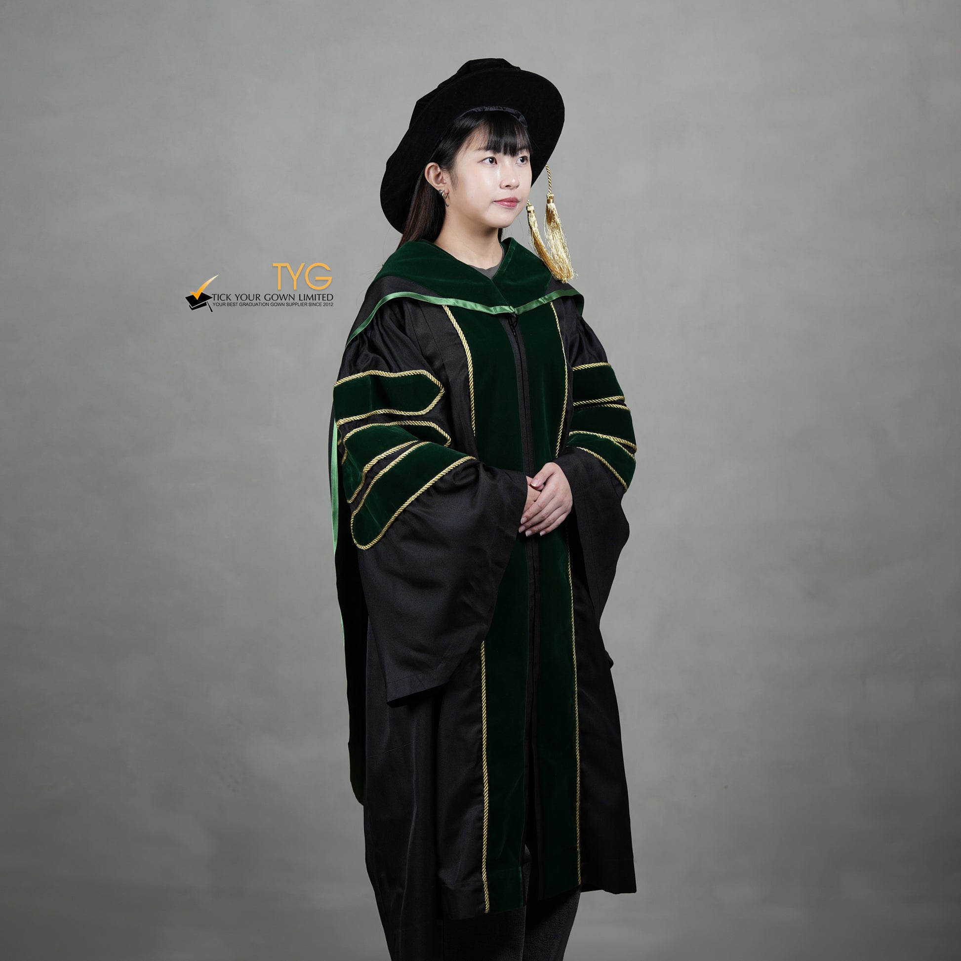 A profile view of a person wearing a PhD graduation gown in black with forest green velvet panels, matching sleeve bars, gold trim, and a matching doctoral bonnet with gold tassel.