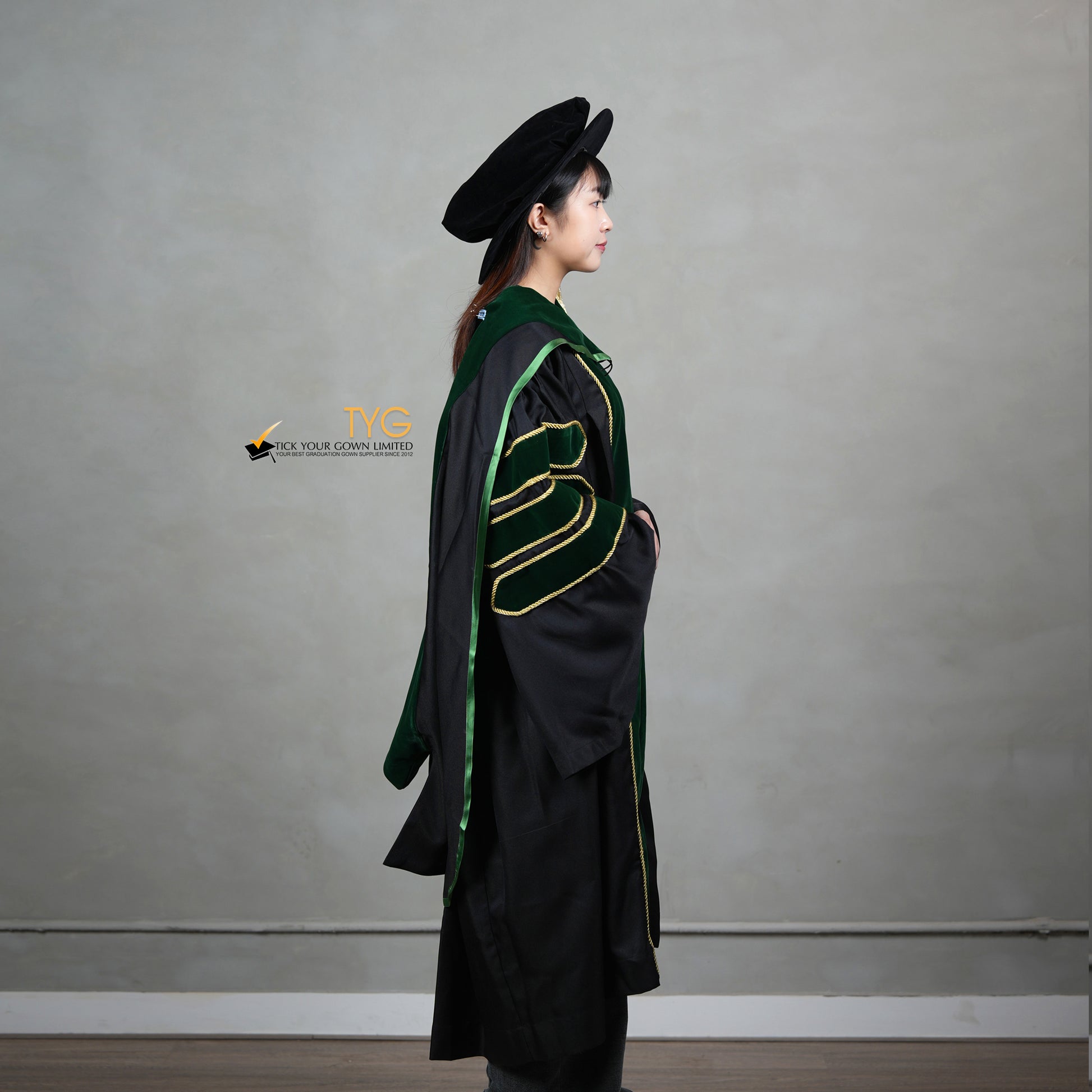 A side view of a person wearing a PhD graduation gown in black with forest green velvet panels, matching sleeve bars, gold trim, and a matching doctoral bonnet with gold tassel.