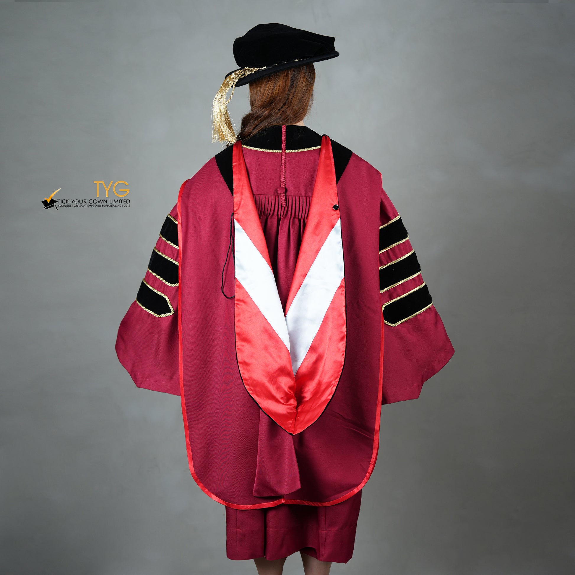 A back view of a person wearing a doctoral graduation gown in maroon with black velvet panels, matching black velvet sleeve bars, gold trim, and a black doctoral bonnet with gold tassel.