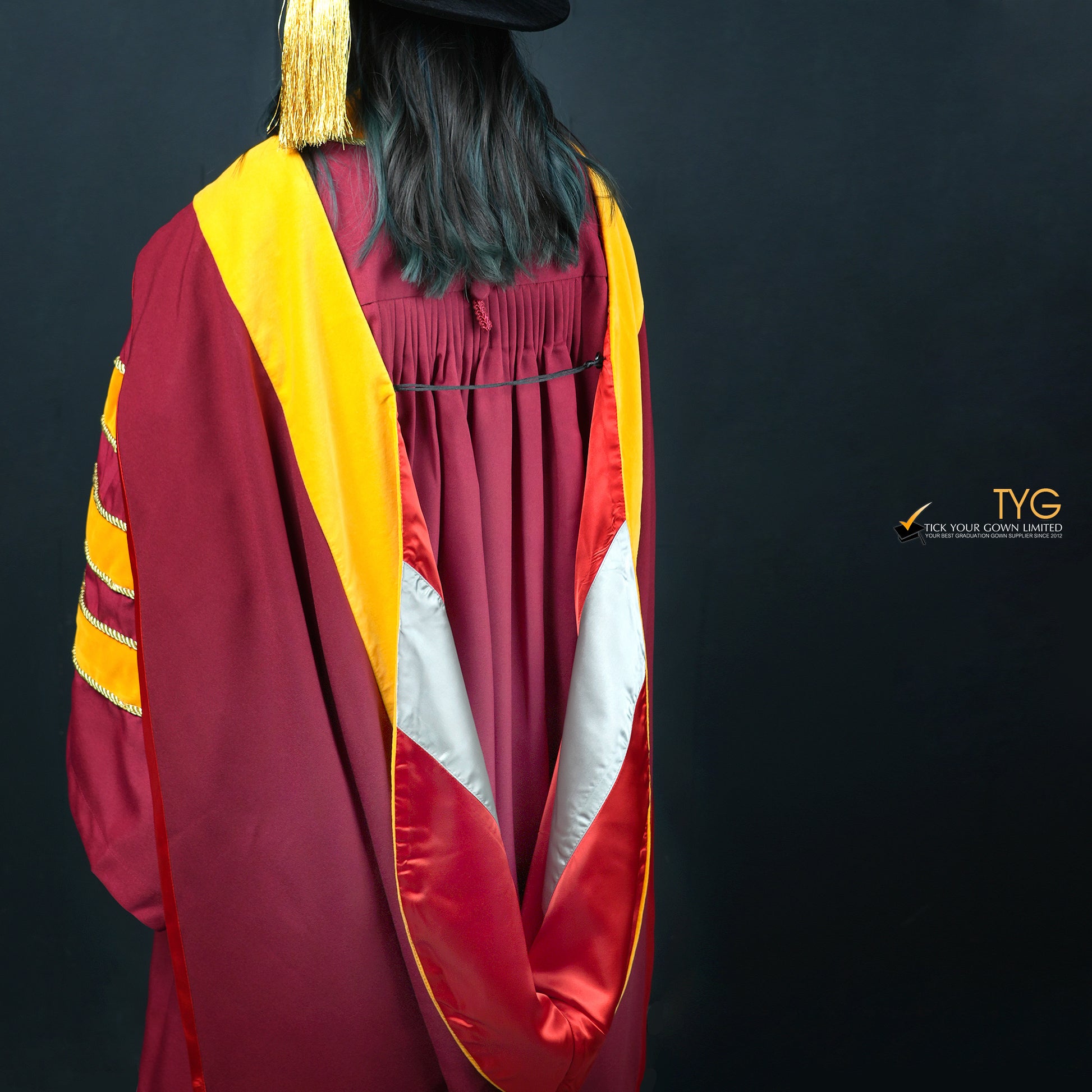 A detailed back profile view of a person wearing a classic PhD graduation gown in maroon with yellow velvet panels, matching sleeve bars, and a black doctoral bonnet with gold tassel