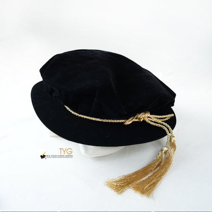 Black velvet doctoral bonnet hat with gold tassels on a white background