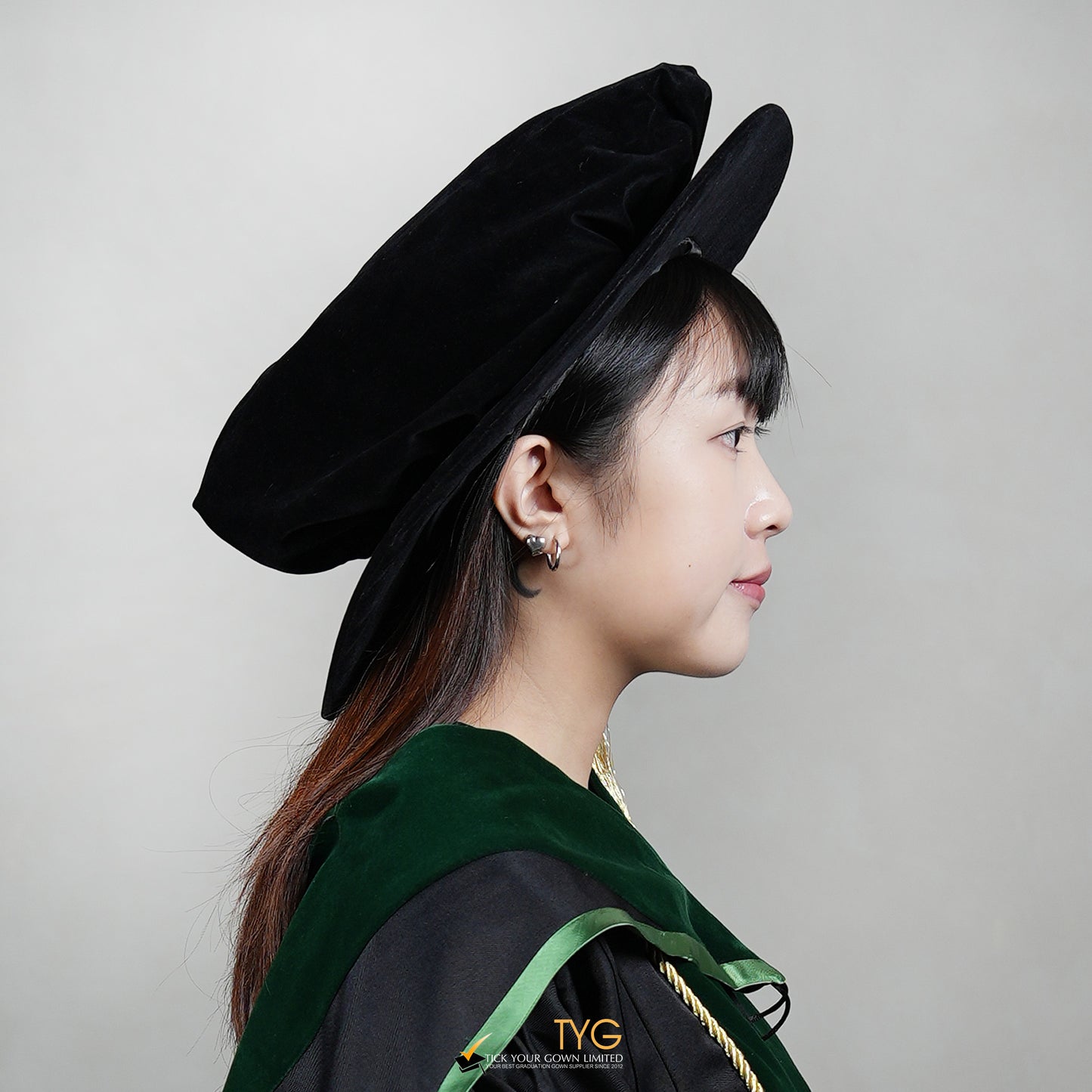 Side view of a person wearing a black doctoral bonnet and gown with a green stole on a plain background