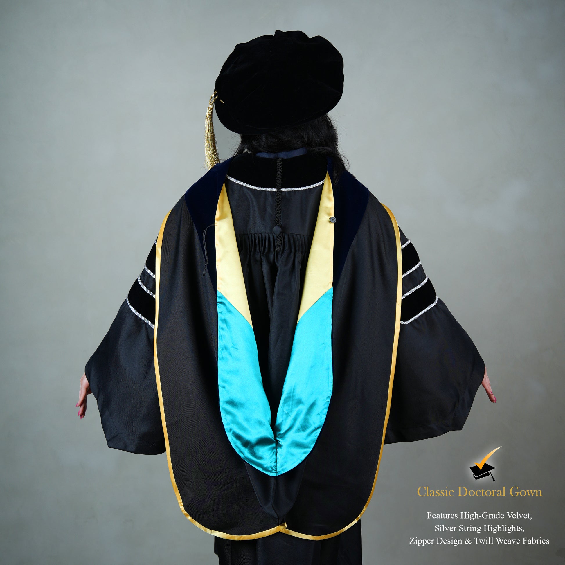 Back view of a person wearing a PhD classic black velvet graduation gown with silver sleeve bars, silver front trim, and a black doctoral bonnet with gold tassel.