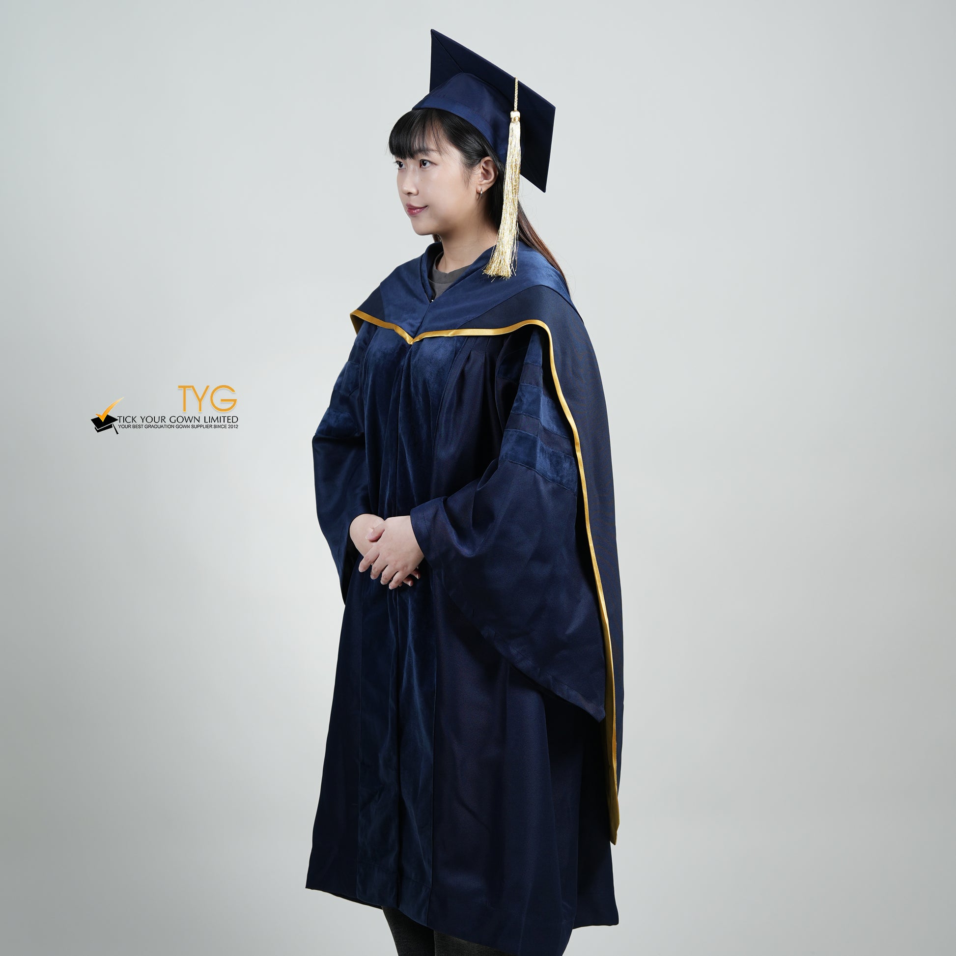 A profile view of a person wearing a PhD classic navy blue velvet graduation gown with yellow lining hood, and a navy blue doctoral bonnet with gold tassel.